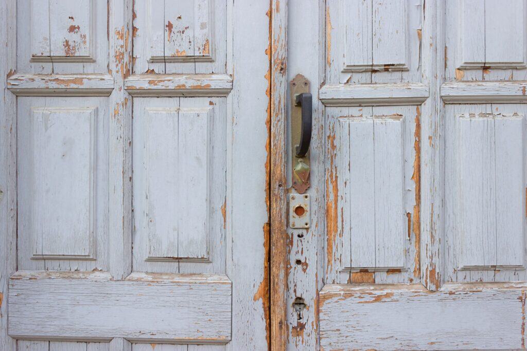 Close-up of an old, weathered wooden door with peeling white paint and a metal handle.
