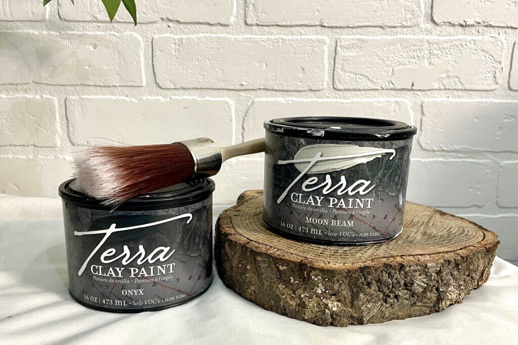 Two cans of Terra Clay Paint, labeled Onyx and Moon Beam, sit on a wooden slab; a paintbrush rests on one can, with a white brick wall in the background.