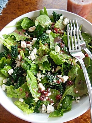 A bowl of mixed green salad topped with crumbled feta cheese and dark vinaigrette, with a fork and knife resting on the side.
