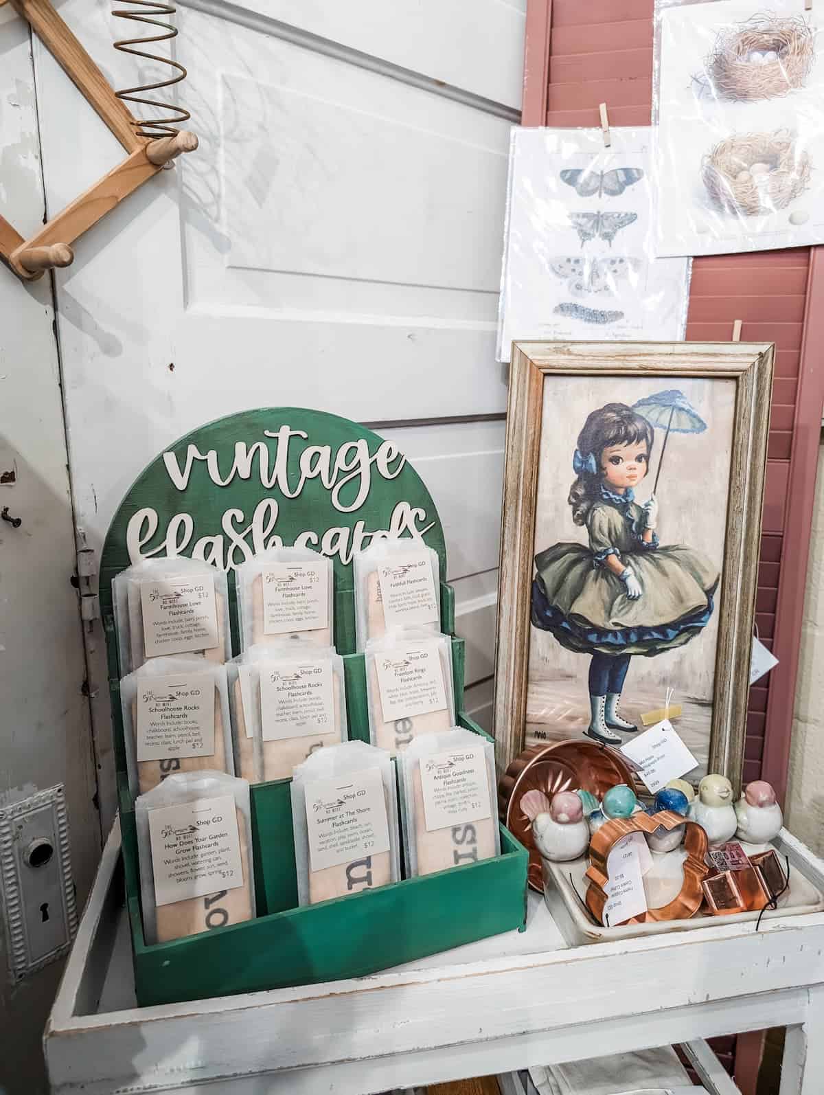 A display with packets labeled “vintage flashcards,” a framed illustration of a girl in period clothing, decorative ornaments, and a wall art print in a vintage shop setting.