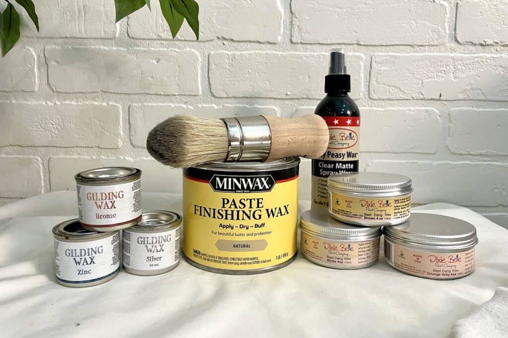 A variety of furniture waxes, a large paintbrush, and a clear matte spray are arranged on a white surface against a white brick wall background.