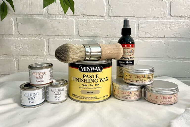 A variety of furniture waxes, a large paintbrush, and a clear matte spray are arranged on a white surface against a white brick wall background.