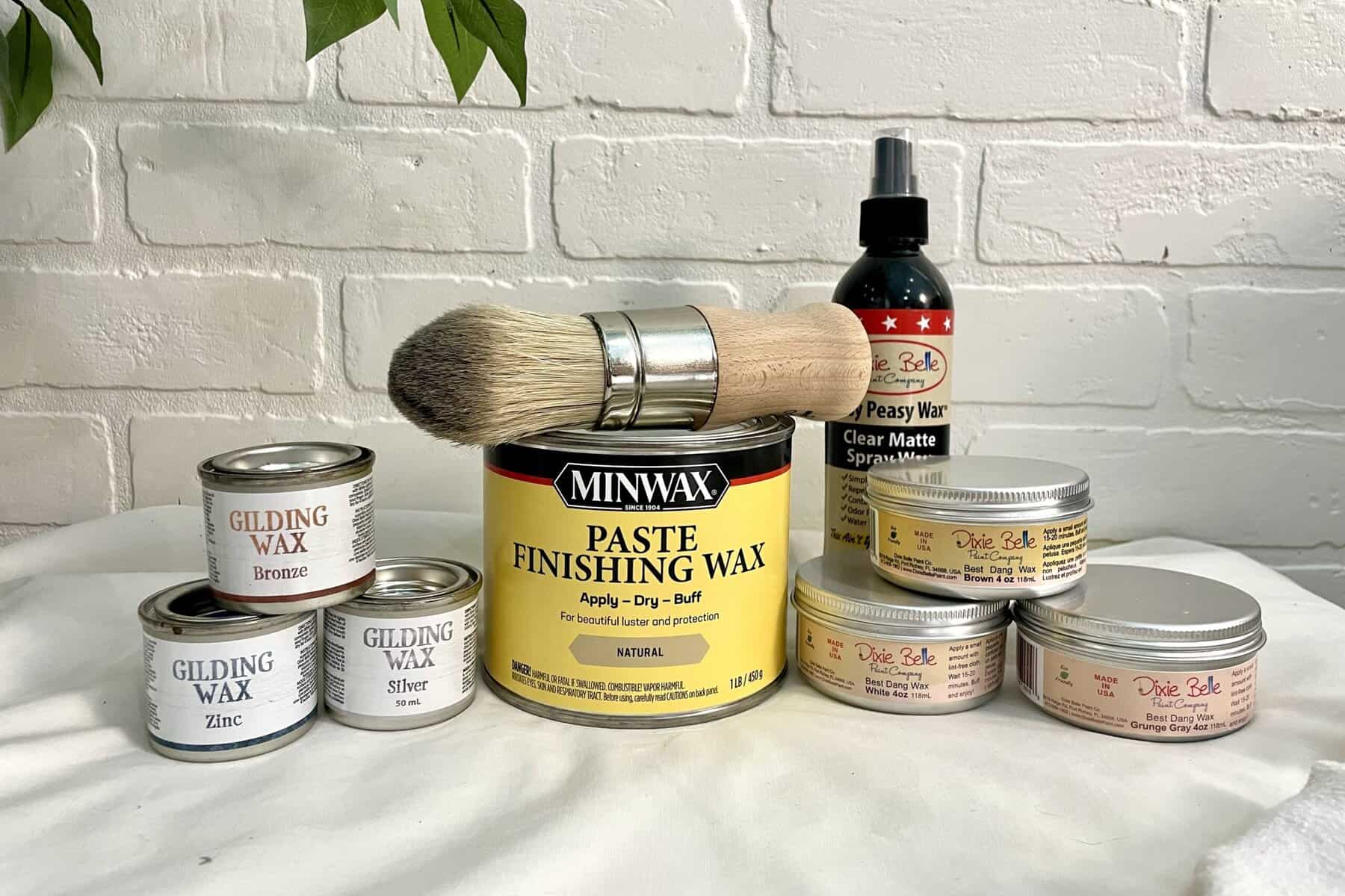 A variety of furniture waxes, a large paintbrush, and a clear matte spray are arranged on a white surface against a white brick wall background.