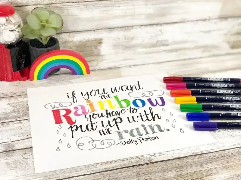 A colorful hand-lettered quote reads "If you want the rainbow, you have to put up with the rain. - Dolly Parton," surrounded by markers, a rainbow, and a plant.