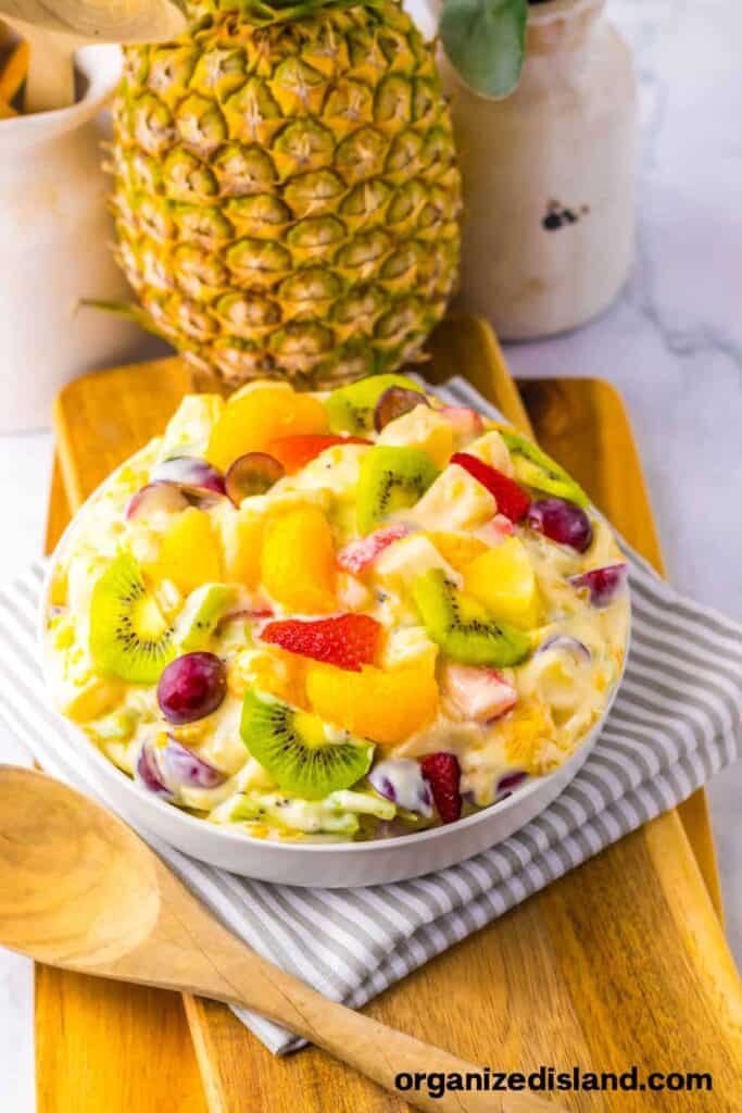 A bowl of creamy fruit salad with kiwi, grapes, strawberries, orange segments, and pineapple, placed on a wooden tray with a whole pineapple in the background.
