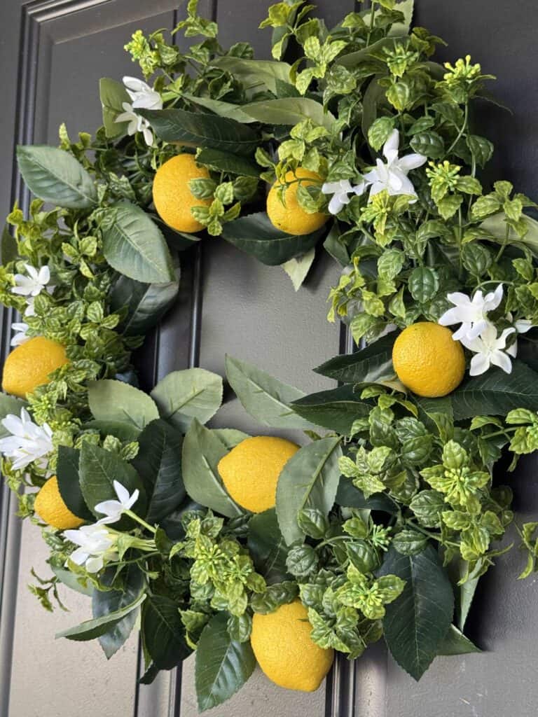 A wreath made of green leaves, white flowers, and artificial yellow lemons hangs on a dark-colored door.
