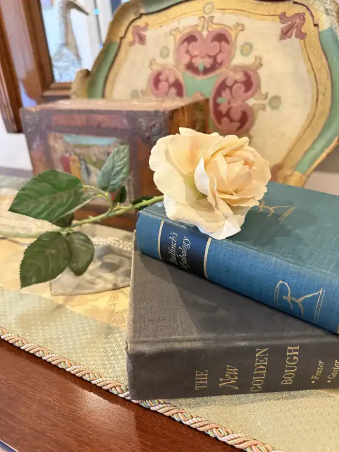 A pale artificial rose rests on two stacked vintage books, one blue and one gray, on a wooden surface with decorative items in the background.