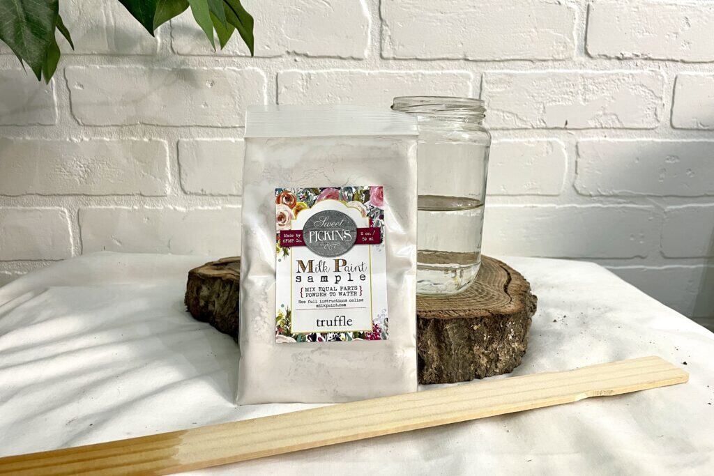 A bag of truffle-colored milk paint, a clear glass jar of water, and a wooden stick are arranged on a white surface with a tree slice and white brick background.
