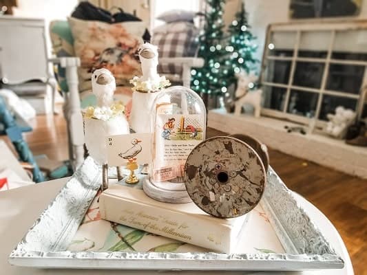 A decorative tray holds a book, a vintage spool, two bird figurines, and a glass dome with cards, set in a cozy room with a Christmas tree and fireplace in the background.