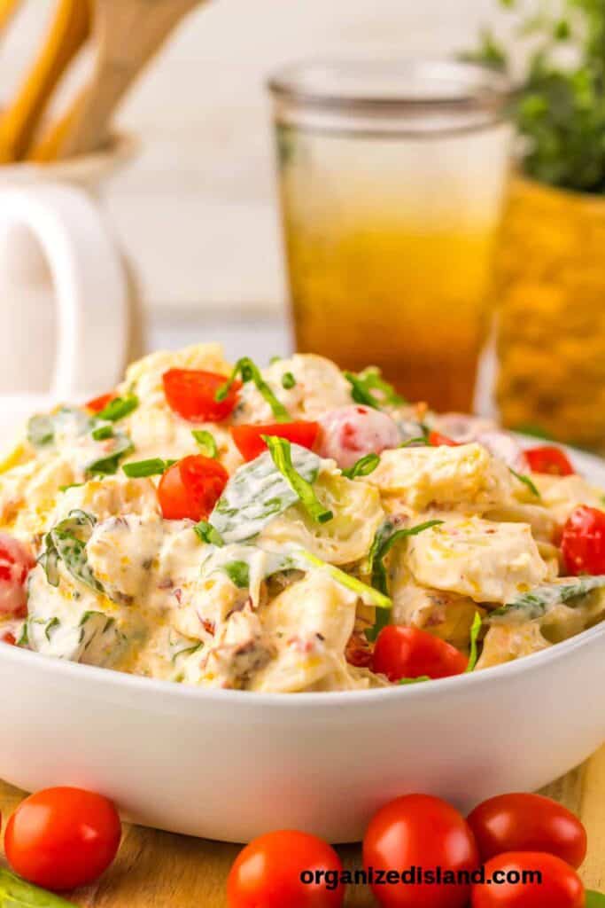 Bowl of creamy pasta salad with cherry tomatoes, spinach, and herbs, placed on a table near a glass of iced tea.