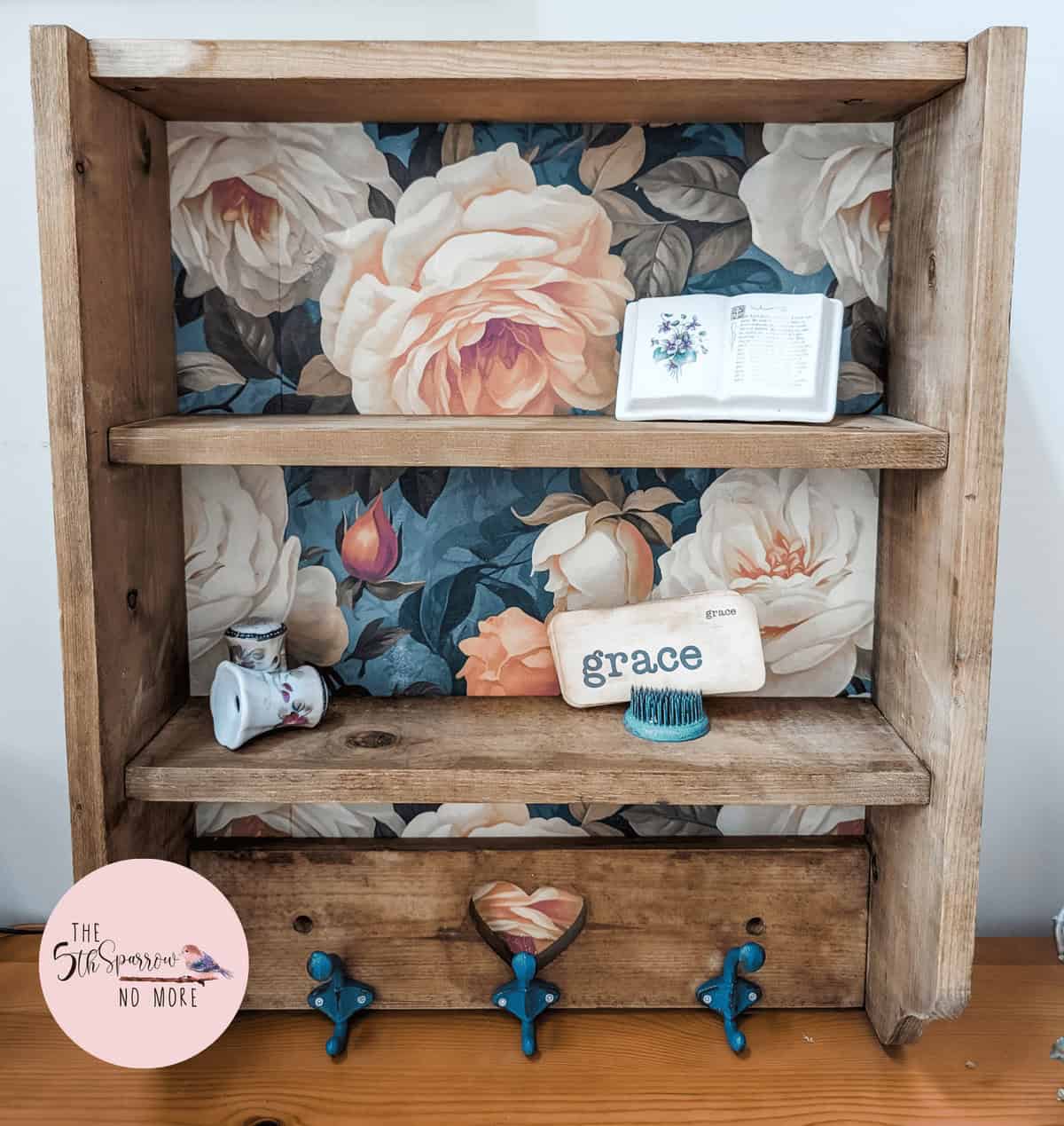 Wooden shelf with floral wallpaper backing, holding a teacup, a plaque reading "grace", a decorative brush, and two small plaques; three blue hooks and a drawer at the bottom.
