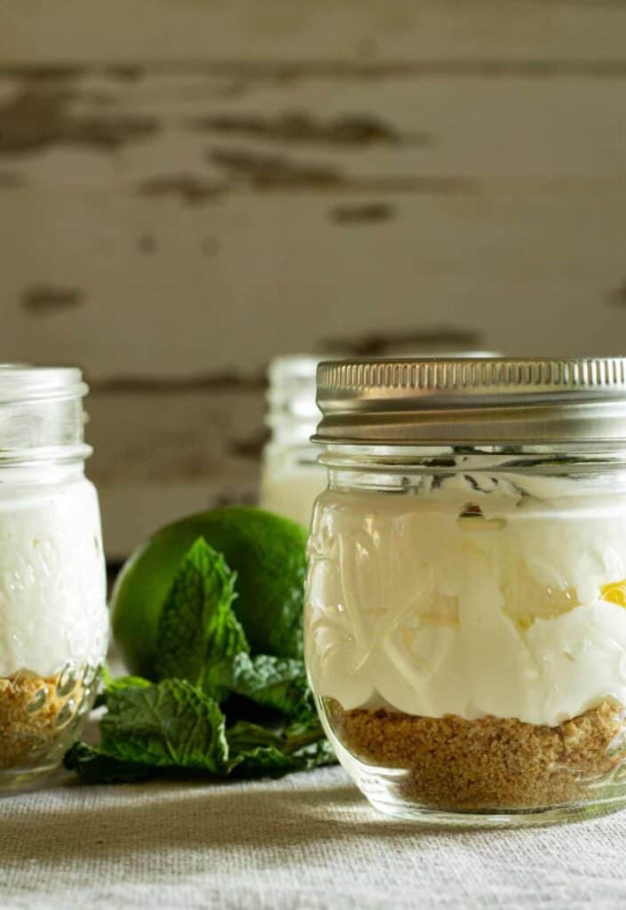Three glass jars containing layered desserts with a crumb base and creamy topping, placed on a table with fresh mint leaves and a lime in the background.