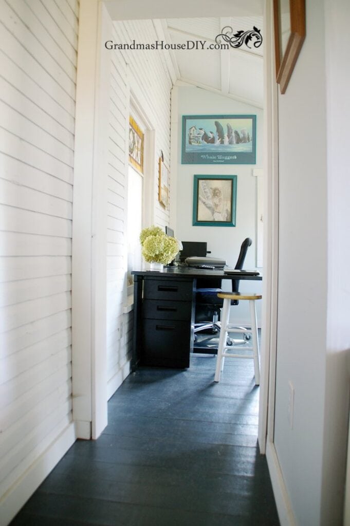 A small home office with a black desk, computer, white chair, and framed pictures on white shiplap walls; a vase of flowers sits on the desk.