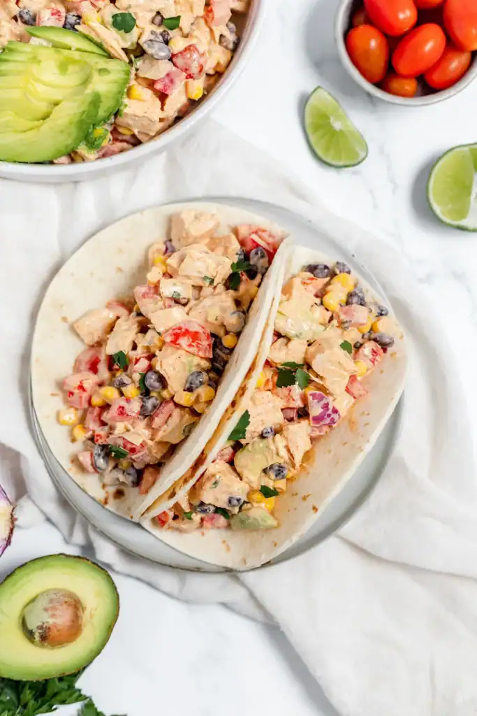 Two tortillas filled with a mixture of chicken, black beans, corn, red onion, and red pepper are served on a plate. Avocado slices, lime wedges, and grape tomatoes are nearby.