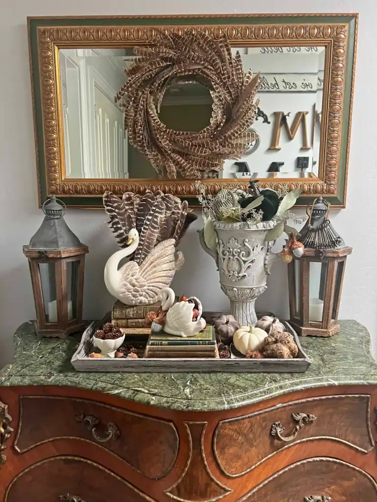 A decorative arrangement on a marble-topped dresser with swan figurines, pumpkins, pinecones, lanterns, a vase with foliage, and a mirror with a wreath above.