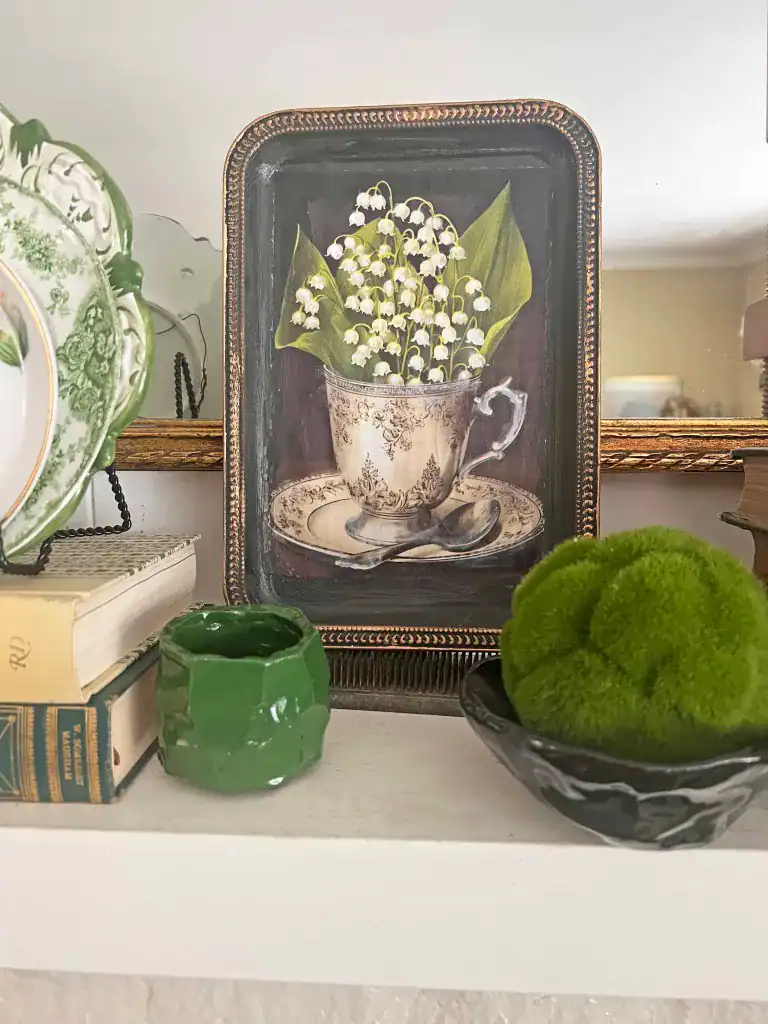 A framed painting of lilies of the valley in a teacup is displayed on a white shelf beside books, a green vase, a plate, and a small bowl with green moss.