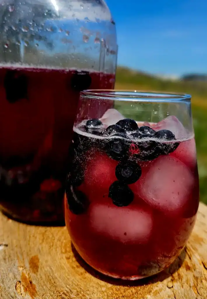 A glass of purple iced drink with blueberries and ice cubes sits on a wooden surface, with a larger jar of the same beverage in the background.