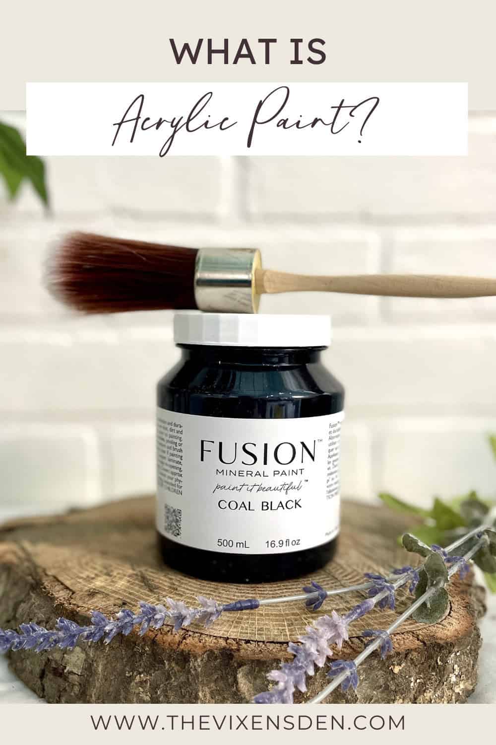 A jar of Fusion Mineral Paint in Coal Black with a paintbrush resting on top, displayed on a wood slice with dried flowers, against a white brick background.