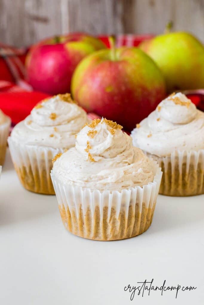 Three cupcakes with light brown frosting are displayed in front of several red and green apples on a white surface.