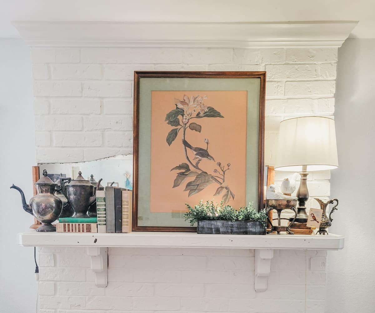 A white mantel holds a framed botanical print, vintage teapots, stacked books, a lamp, and a small green plant against a white brick wall.