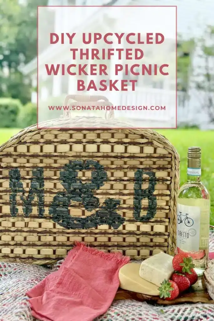 Wicker picnic basket with "M & B" painted on it, wine, strawberries, and a plate on a blanket outdoors. Text overlay reads "DIY Upcycled Thrifted Wicker Picnic Basket.