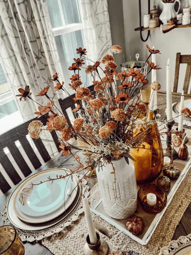 A dining table set with plates, candles, and a white vase of orange flowers, decorated with amber glass bottles and small pumpkins on a woven placemat.