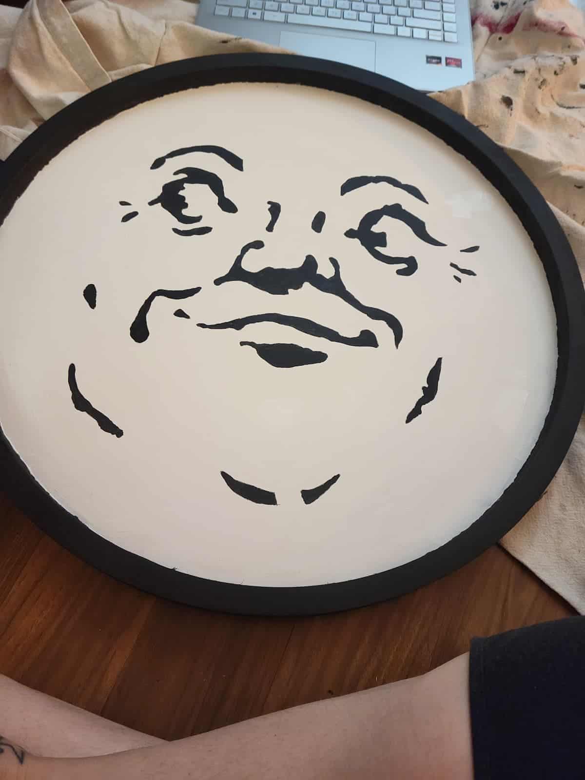 A round object painted with a black outline and a stylized face, resembling the moon, is placed on a wooden floor near an open laptop and a person's arm.