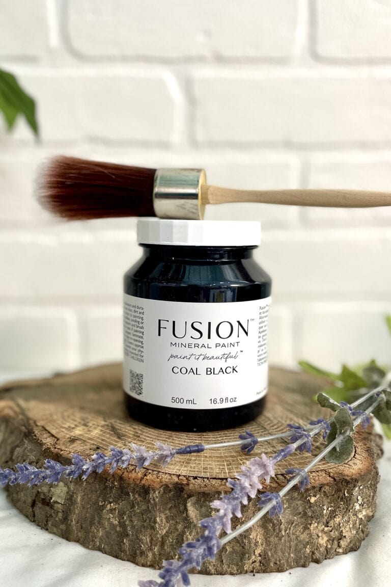 A jar of Fusion Mineral Paint in coal black with a paintbrush resting on top, placed on a wooden surface with lavender sprigs, against a white brick background.