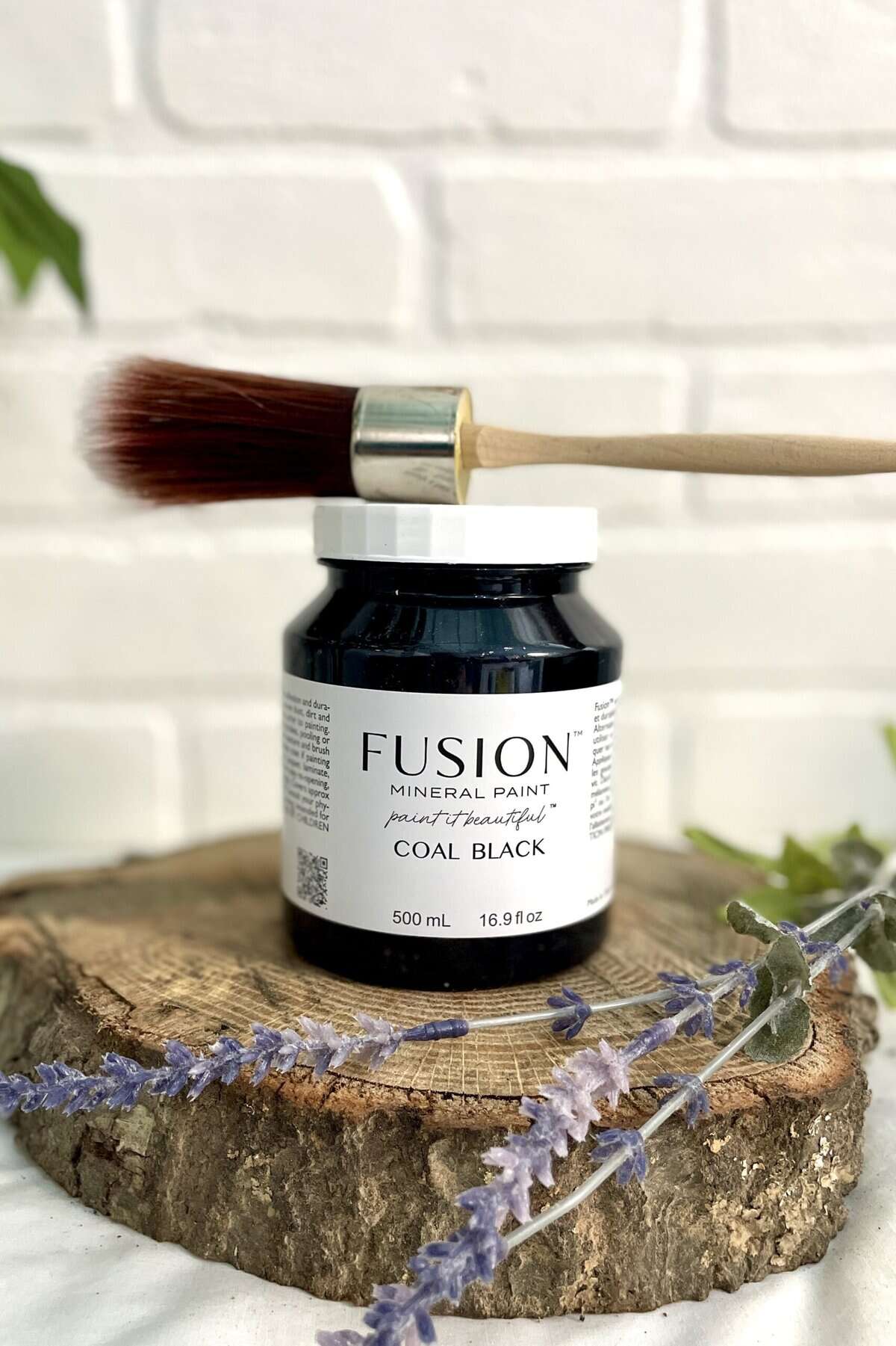 A jar of Fusion Mineral Paint in coal black with a paintbrush resting on top, placed on a wooden surface with lavender sprigs, against a white brick background.