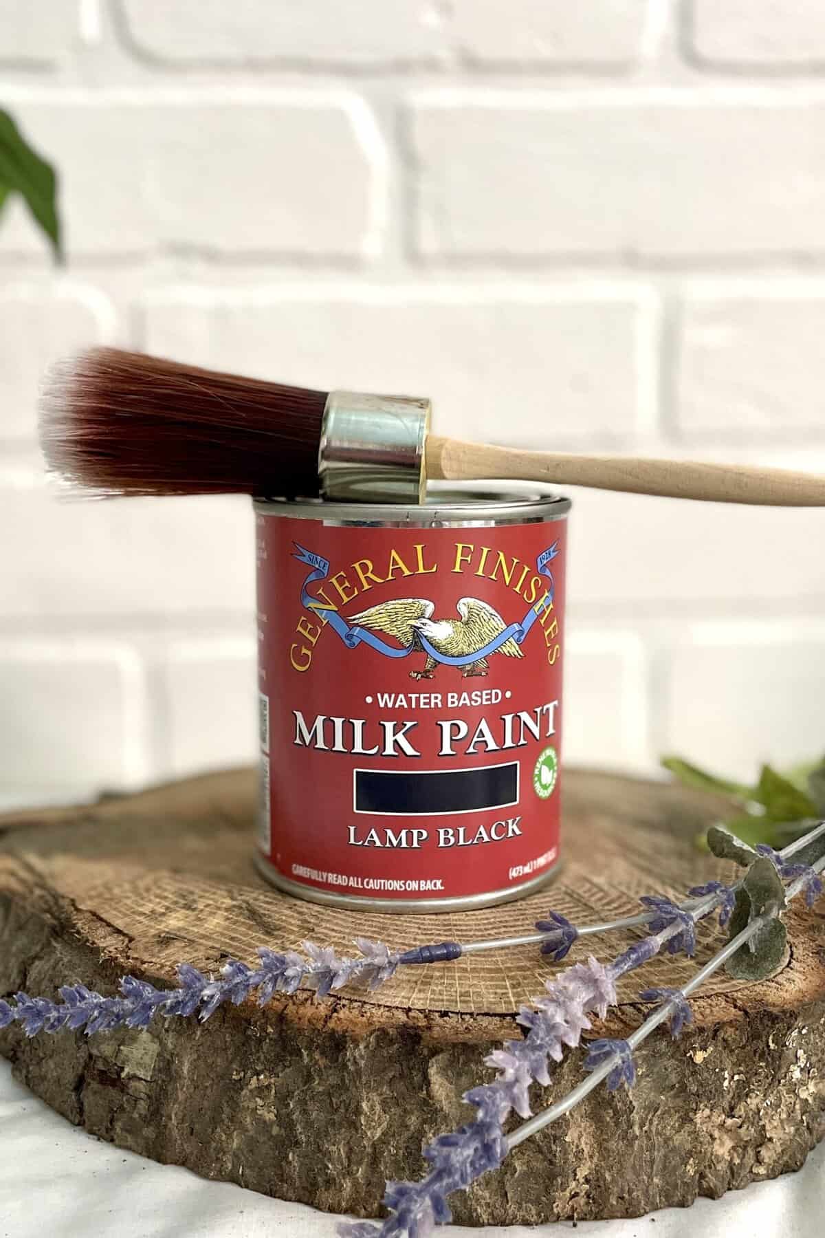 A can of General Finishes Lamp Black milk paint with a paintbrush resting on top, placed on a wooden surface against a white brick background.
