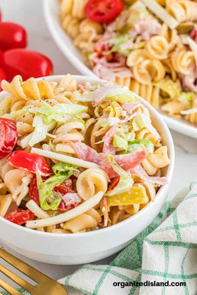 A bowl of pasta salad with rotini, cherry tomatoes, lettuce, sliced deli meat, and cheese, served next to a dish of the same salad.