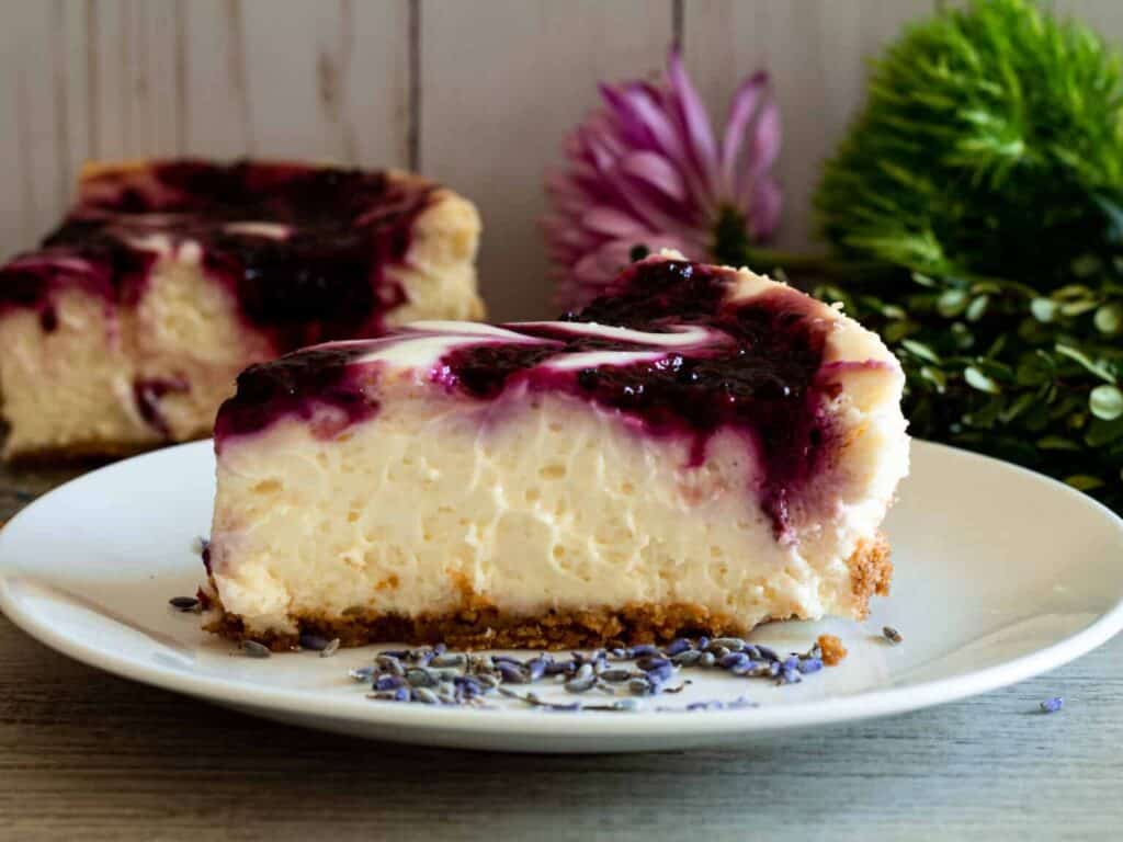 A slice of cheesecake with a graham cracker crust and dark berry topping on a white plate, with flowers and greenery in the background.