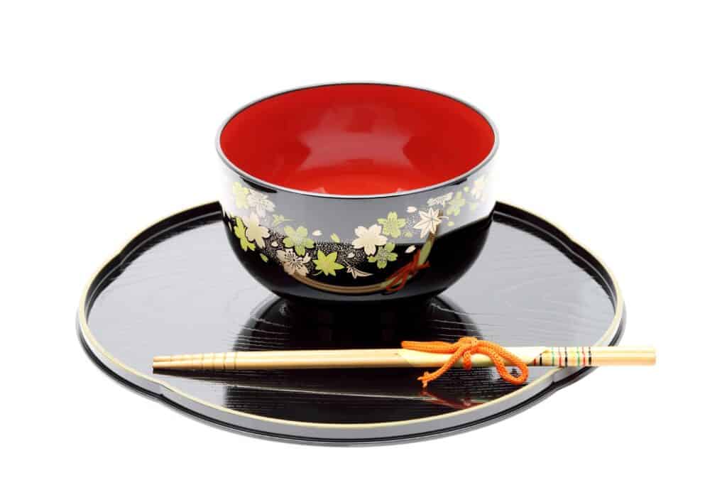 A black and red lacquered bowl with floral designs sits on a matching tray, accompanied by decorated chopsticks tied with an orange cord.