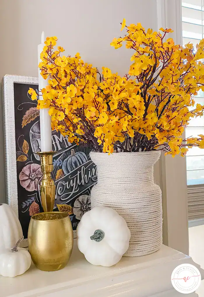 White vase with yellow flowers, a gold candlestick with white candle, a gold cup, two white pumpkins, and a chalkboard sign arranged on a white surface.