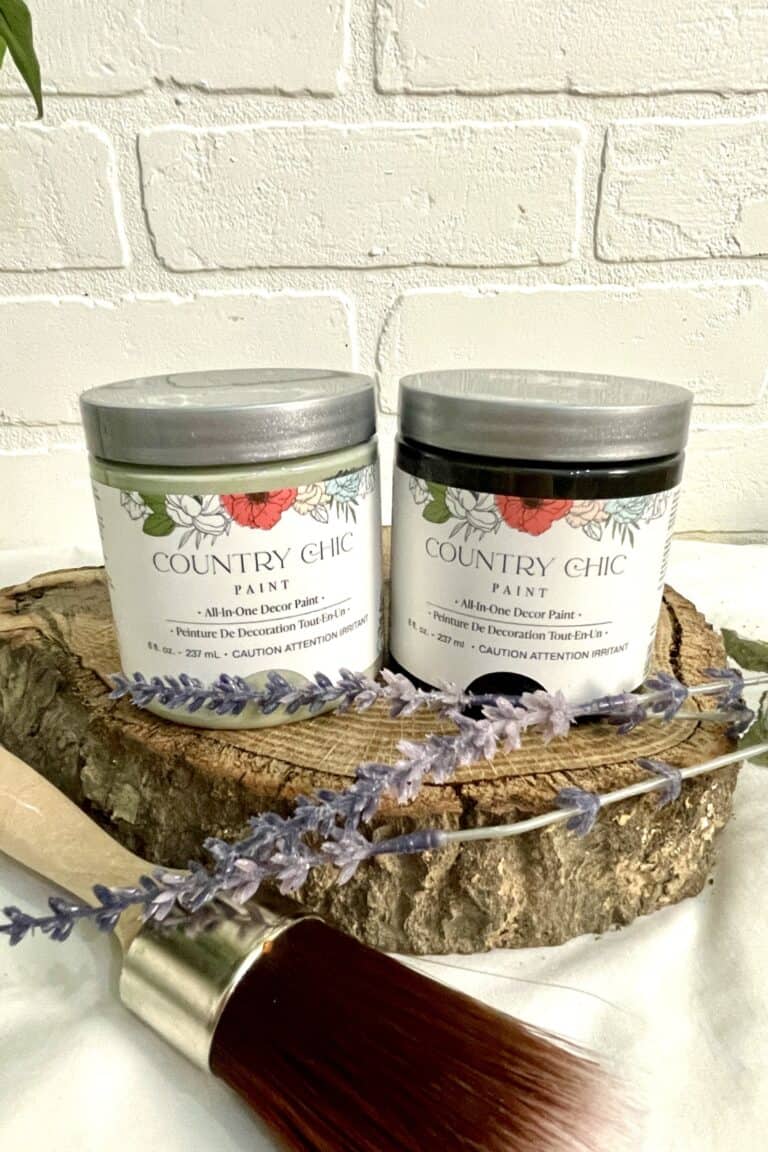 Two containers of Country Chic Paint sit on a wooden slab with a paintbrush and sprigs of lavender in front, against a white brick wall background.