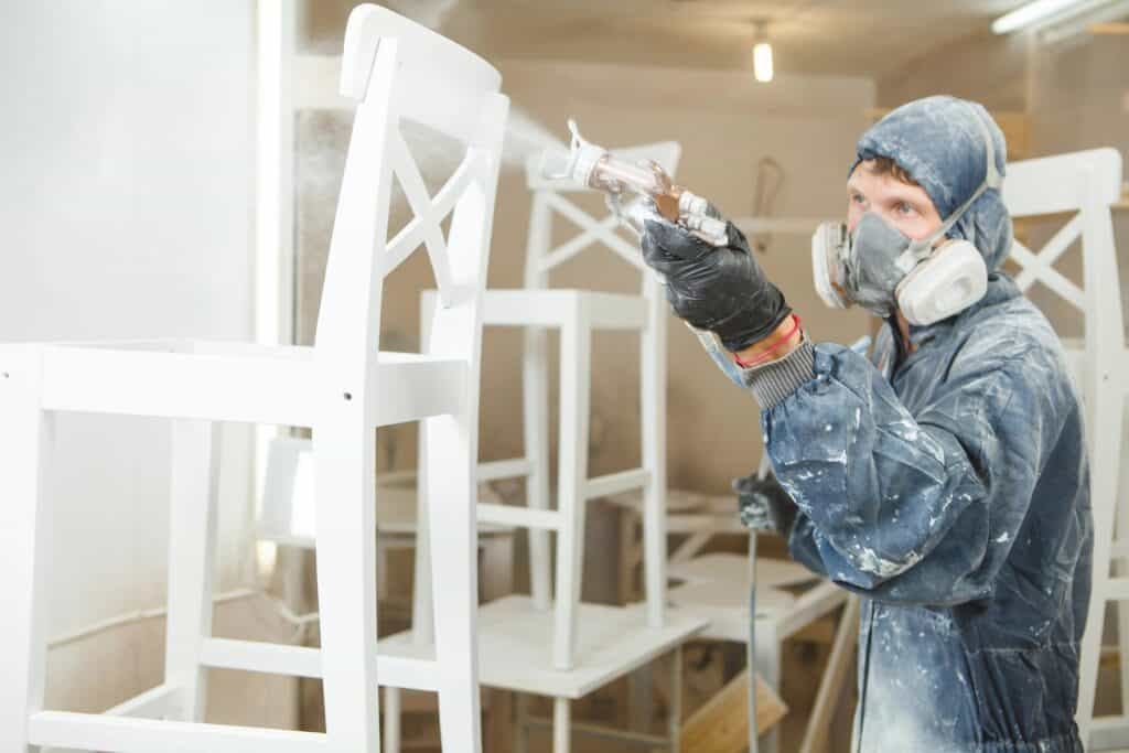 A person in protective gear spray paints a wooden chair white in a workshop.