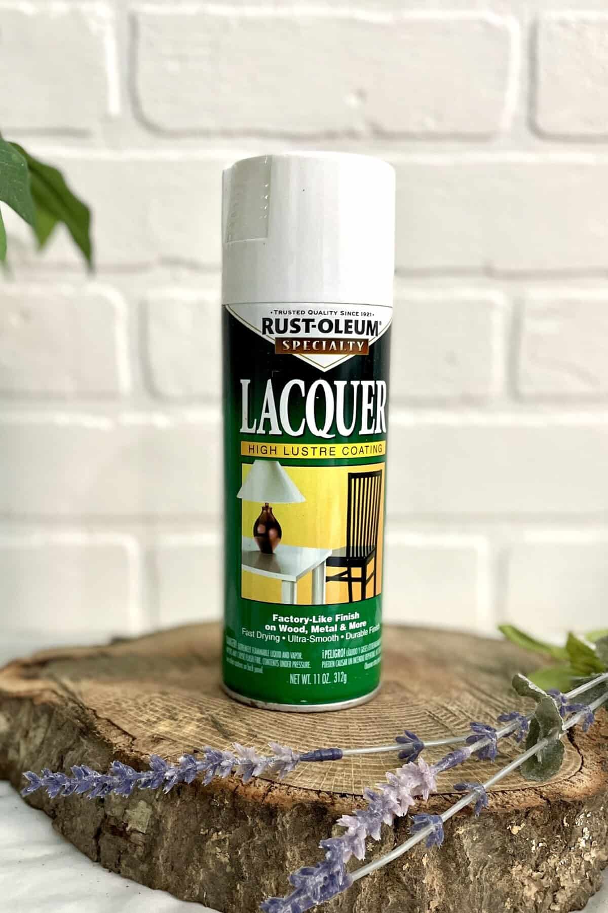 A can of Rust-Oleum Specialty Lacquer spray sits on a wooden surface with some lavender flowers in front, against a white brick background.