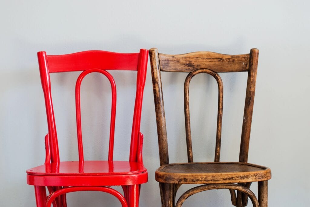 A red wooden chair and a weathered brown wooden chair are positioned side by side against a plain light gray wall.