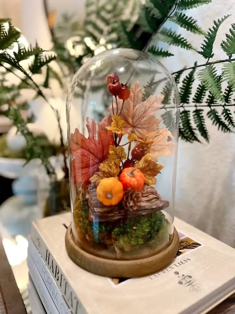 A glass cloche displays artificial autumn leaves, berries, and small pumpkins on a bed of moss, placed on stacked books with fern leaves in the background.