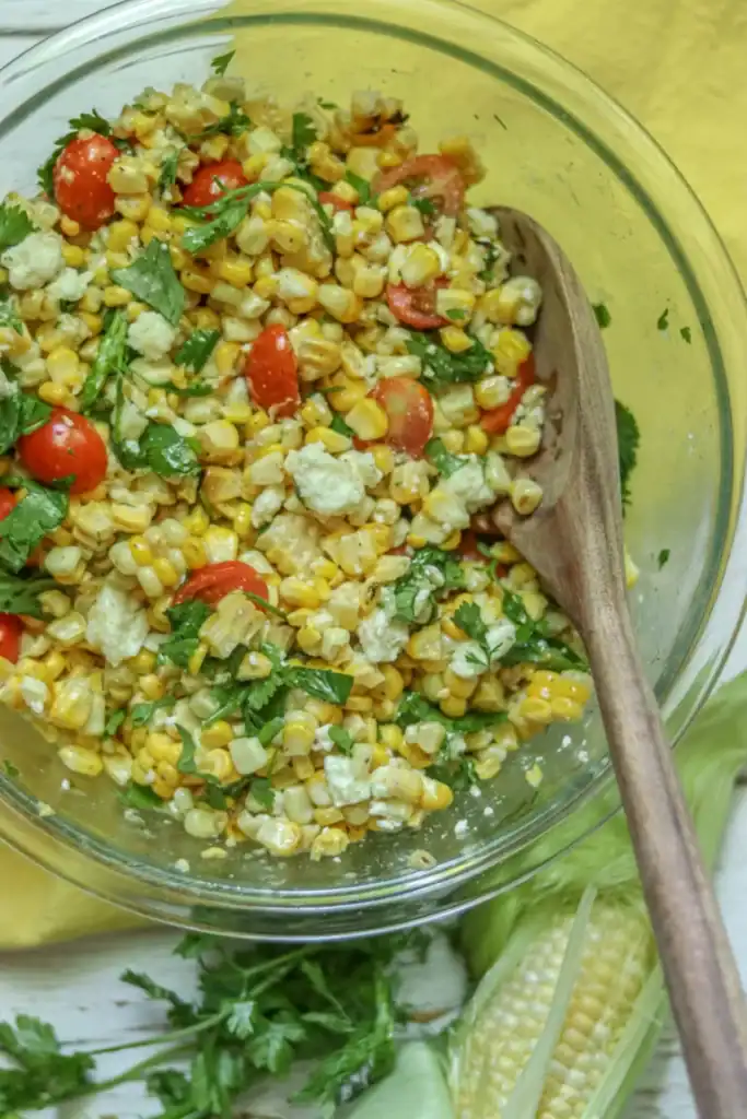 A glass bowl of corn salad with tomatoes, feta cheese, and cilantro, mixed with a wooden spoon. An ear of corn and some cilantro are on the table beside the bowl.