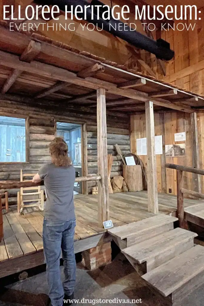 A person stands inside the Elloree Heritage Museum, viewing a rustic wooden cabin exhibit with informational signs on the walls.