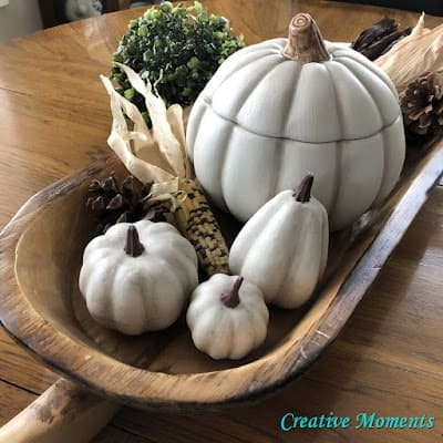 Wooden tray with white decorative pumpkins, a green faux plant ball, dried corn, pine cones, and wooden table surface underneath. "Creative Moments" text is in the corner.