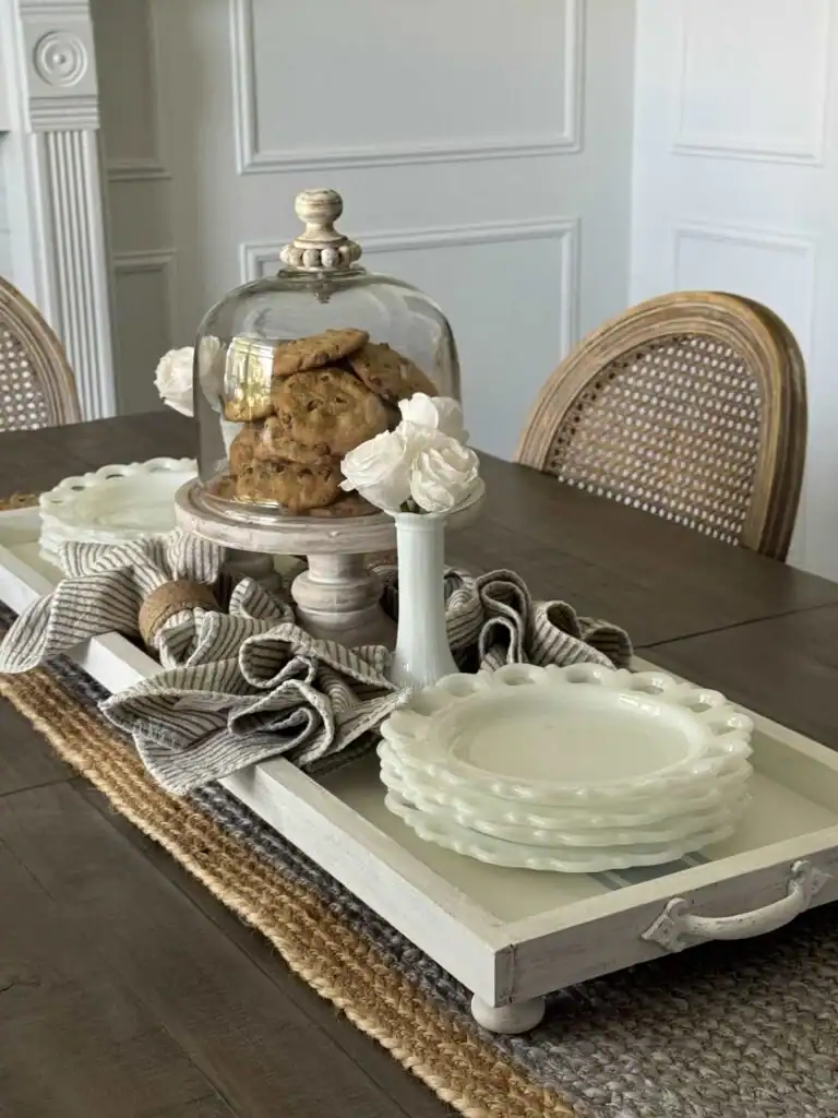 A wooden tray on a dining table holds stacked white plates, striped napkins, a white vase with flowers, and cookies under a glass dome.