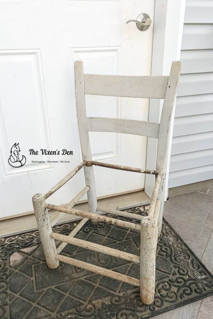 A worn, white wooden chair with missing seat and chipped paint sits on a doormat in front of a closed white door.