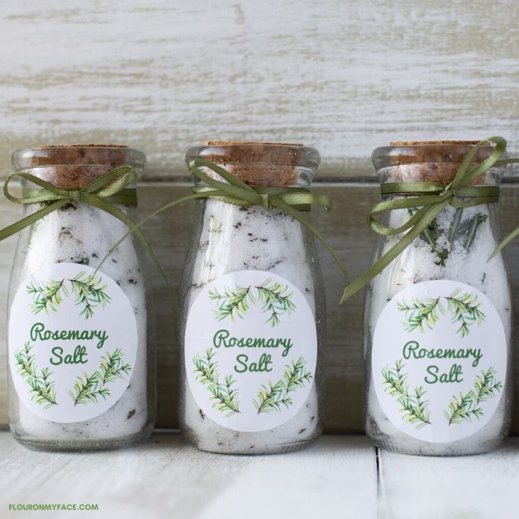 Three glass jars filled with rosemary salt, each sealed with a cork and tied with a green ribbon, are labeled "Rosemary Salt" with a wreath design.