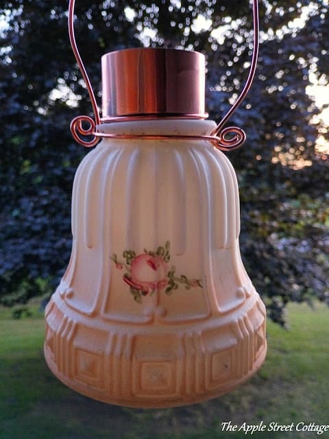A vintage hanging lamp with a frosted glass shade, floral design, and metal top is shown outdoors with trees in the background.