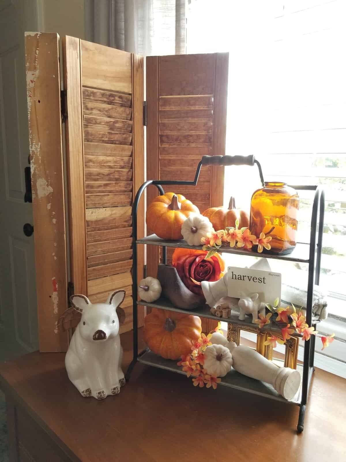 A two-tiered stand with pumpkins, faux flowers, an amber jar, and a “harvest” sign sits on a table next to a ceramic pig, with wooden shutters and sunlight in the background.