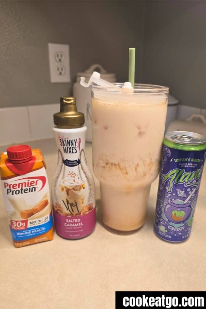 A glass of iced beverage with a straw is placed beside Premier Protein shake, Skinny Mixes salted caramel syrup, and a can of Alani Nu energy drink.