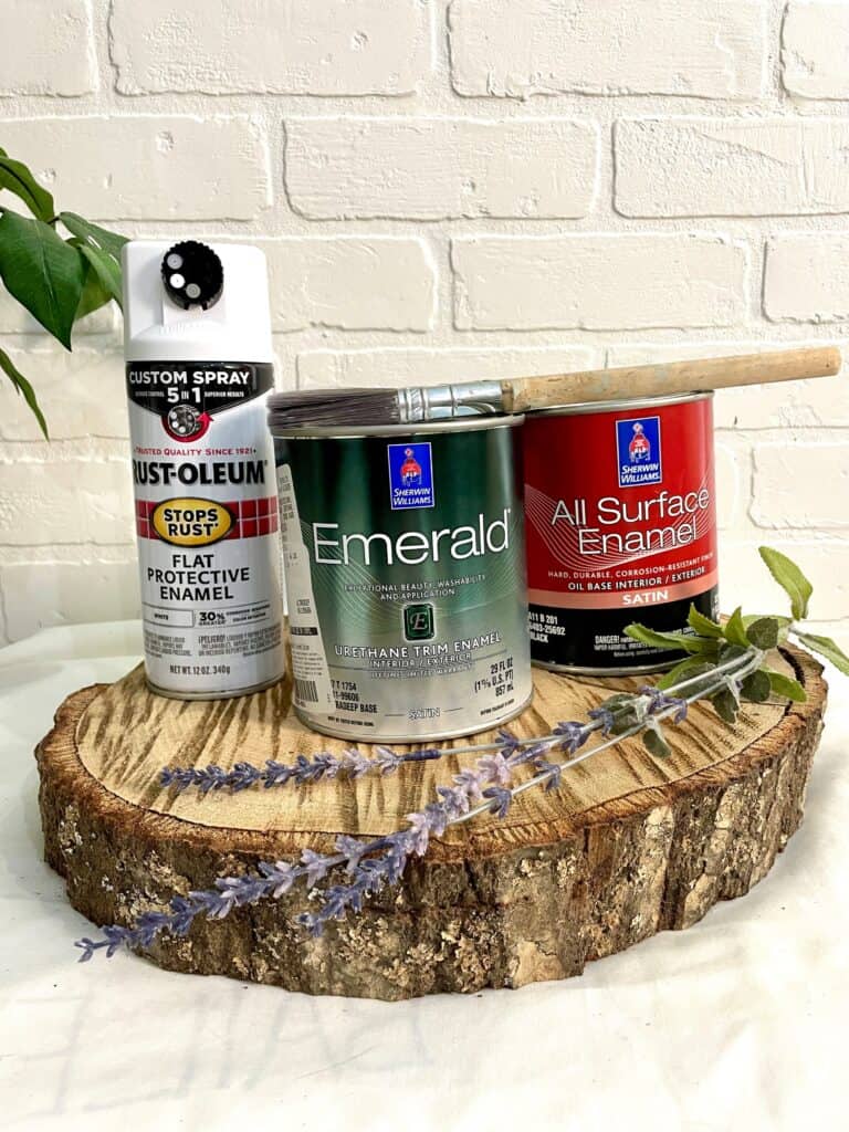 Three cans of paint, including spray and enamel types, sit on a wood slice with a paintbrush, lavender sprig, and green leaves against a white brick wall.
