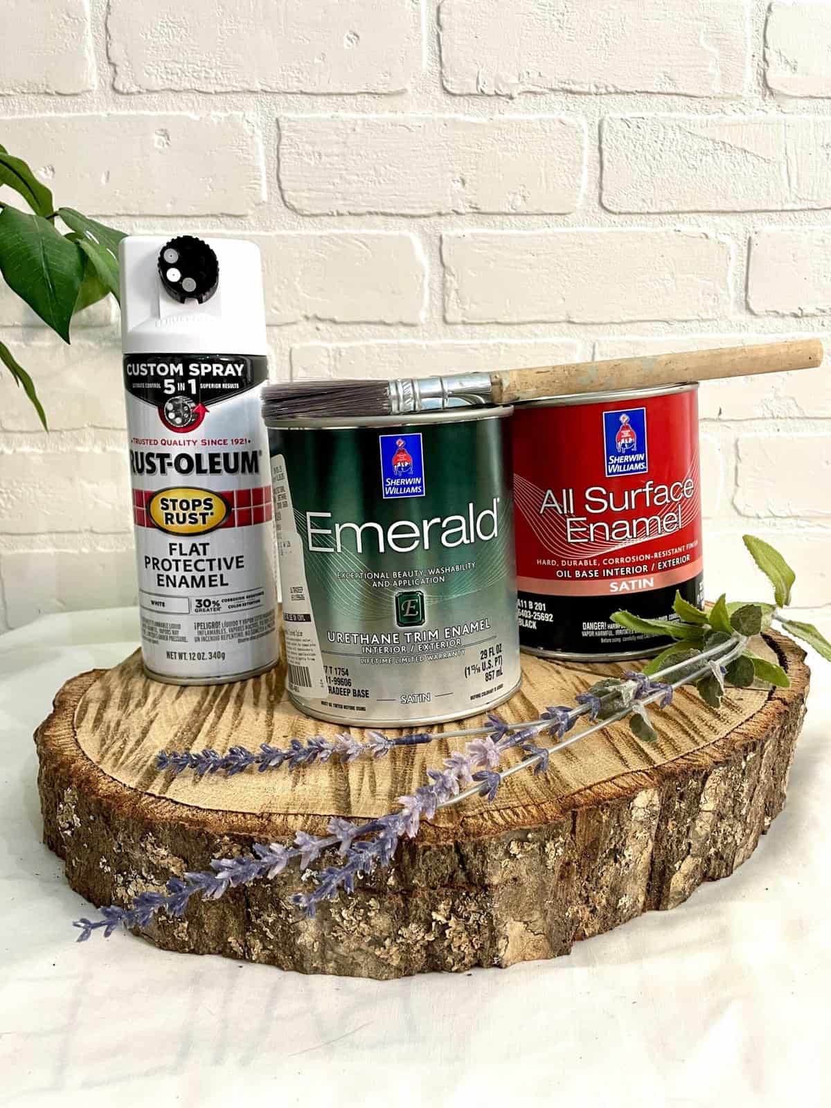 Three cans of paint, including spray and enamel types, sit on a wood slice with a paintbrush, lavender sprig, and green leaves against a white brick wall.